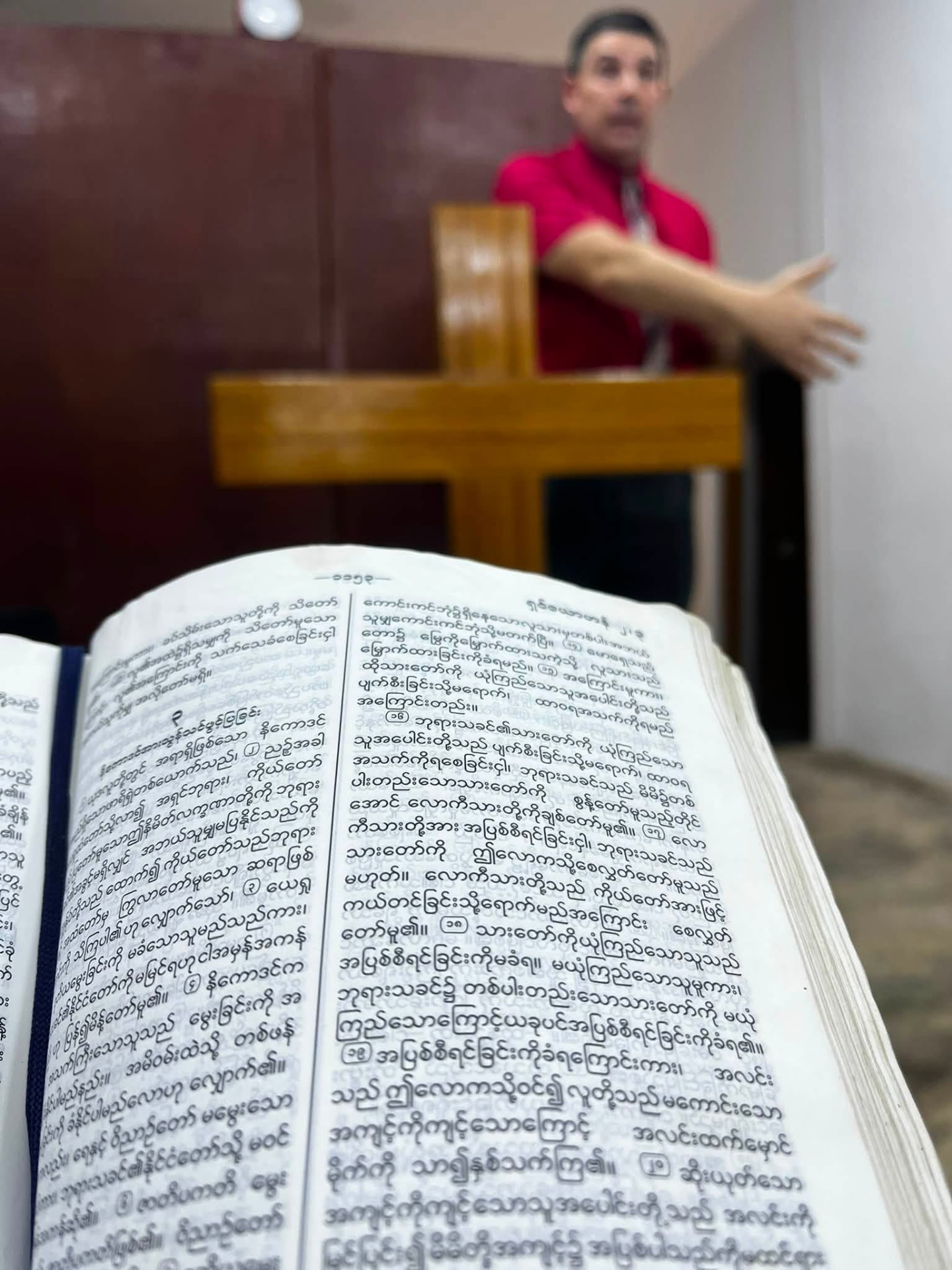 Preaching from the pulpit beside a wooden cross with an open Burmese Bible in the foreground